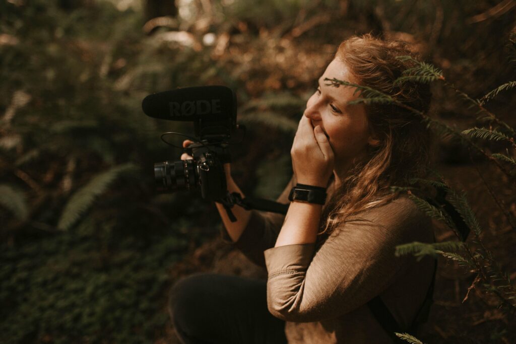 A photographer and videographer is in the redwoods filming a wedding and she is looking at a couple with a hand over her mouth in awe.