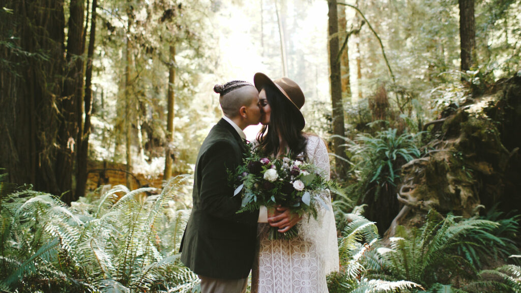 A couple stands in front of a forest of ferns kissing in their wedding clothing in the Redwoods National Park.
