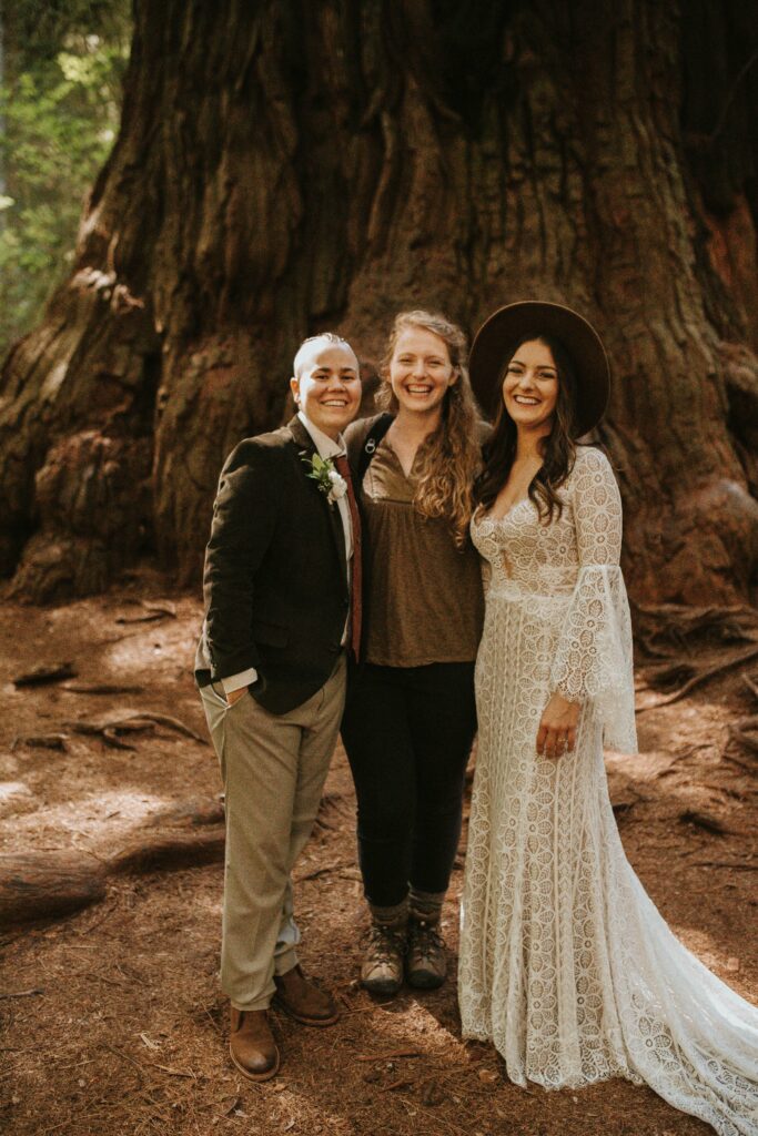 A photographer and videographer stands with their couple smiling at the camera for a portrait during their redwoods wedding..