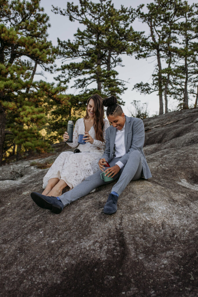 An LGBTQ couple is sitting side by side on a rock to elope in NC. They are making tea and laughing in their wedding clothes. One has a blue suit on and the other a white dress.