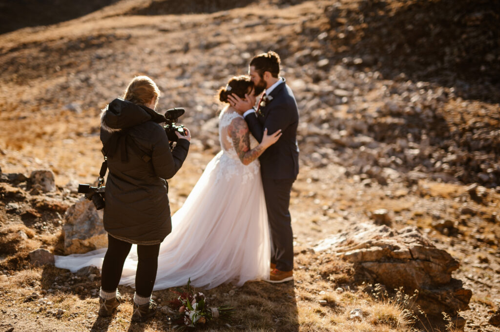 An adventure elopement videographer is filming a couple hugging and the groom is kissing the bride on the head. It is their wedding day and they are outside in nature.