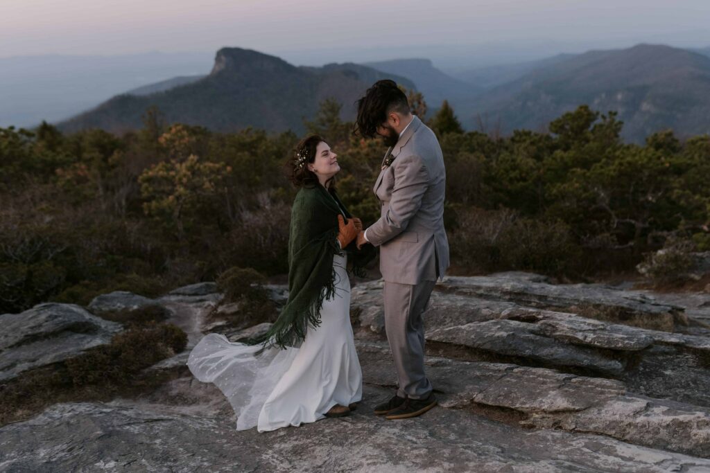 A couple is standing on a mountain at sunrise when the light is still blue and the bride has a blanket on and the groom has a grey suit. It is during their adventure elopement and they have a videographer capturing them.