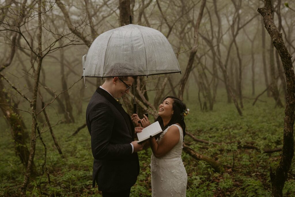 A couple stands in a rainy forest with an umbrella over them during their Asheville elopement. They are dressed in wedding clothes and the groom is reading vows to his bride while she looks at him and laughs.