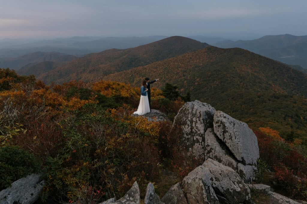 A couple stands on a rock overlooking the mountains during their blue ridge parkway wedding in the middle of fall. It is sunrise and the colors are soft amidst the red and orange leaves. The groom is hugging the bride and pointing into the distance.