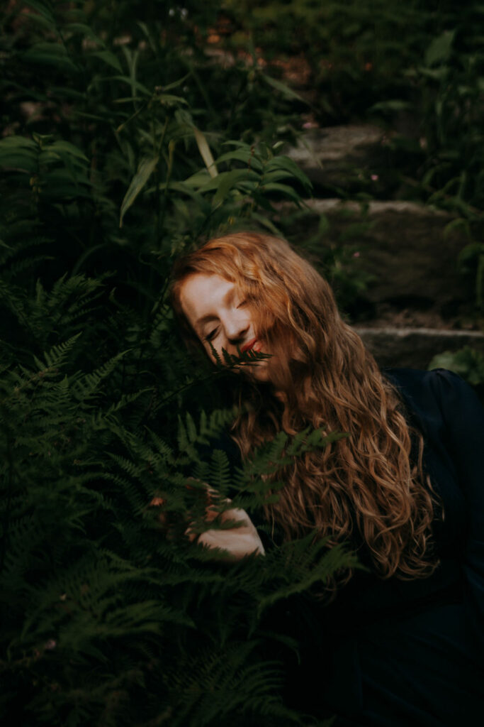 A girl with red curly hair is sitting in a field of ferns smiling. She is an outdoor elopement photographer in Asheville nc.