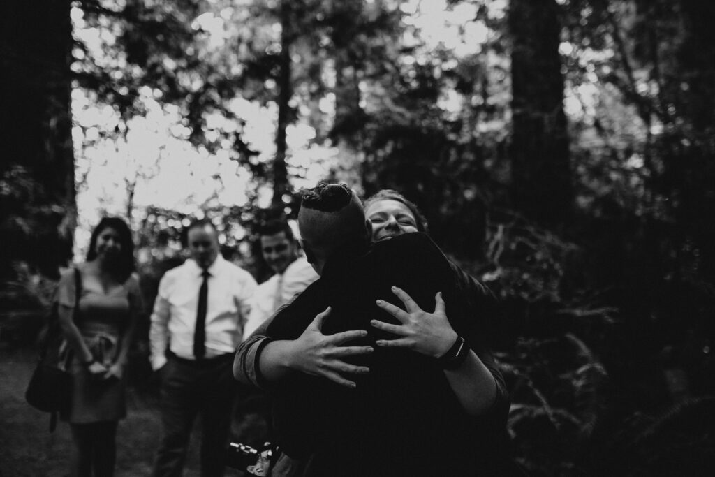 An adventure elopement videographer is hugging the perosn they just filmed get married during their ceremony. The image is black and white and the persons hands are spread wide on the other ones back and they are smiling.