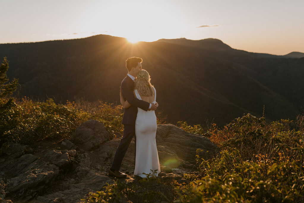 Breanna and Trevor had an elopement that they really had to plan for - they decided to hike up a mountain for sunrise in their wedding clothes! They stand hugging each other as the sun comes over the mountain in the background.