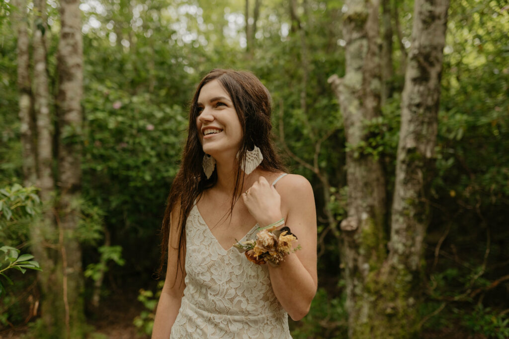 A bride is standing in the forest of North Carolina in her lacy elopement dress smiling up at the sky. She has on a floral wristlet and boho weaving earrings