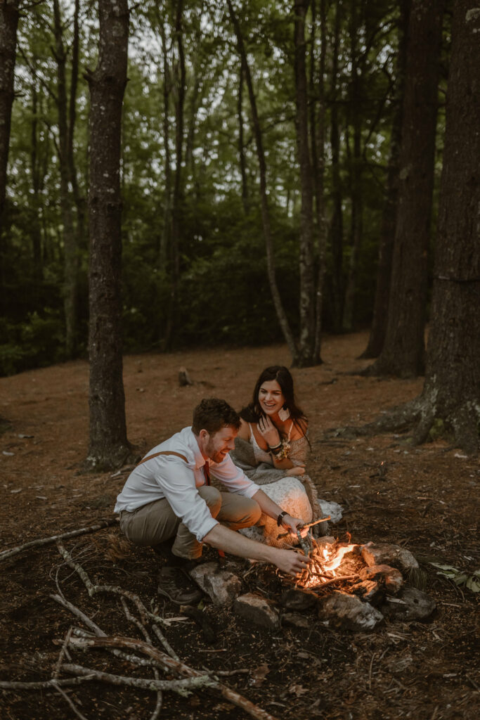 A couple is in a forest at dusk in North Carolina for their elopement day. They are building a fire together, smiling and laughing as the flames pick up and brighten up their faces with warm light.