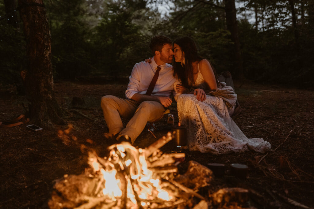 A couple sits in their elopement clothes about to kiss by the campfire. They are in a forest in North Carolina and it is dusk.