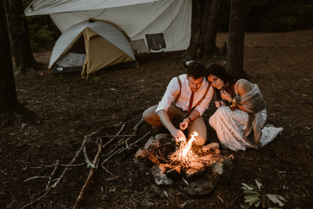 A couple is making a campfire during their elopement in the forest of NC surrounded by pine nettles and trees. Their tent is behind them with a tarp over it to protect from rain.