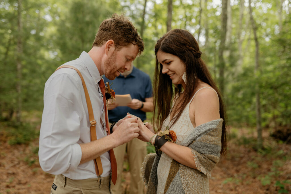 A groom is putting a ring on his brides finger during their elopement in a North Carolina forest elopement.