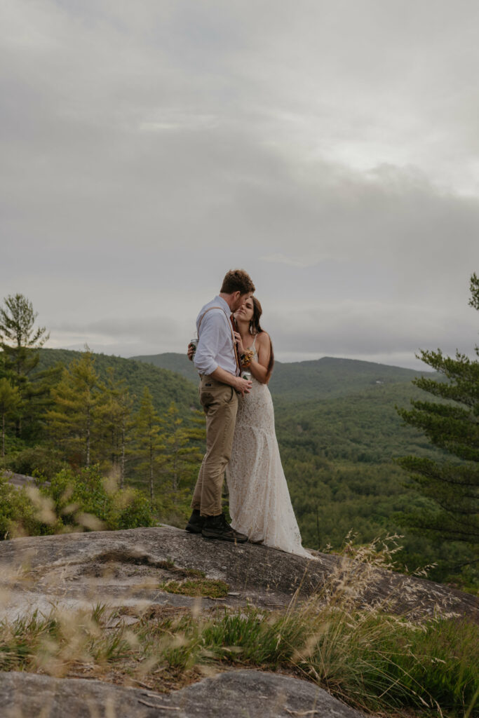 A couple is standing on a rock in North Carolina in their elopement clothing cuddled up. The sky is cloudy and grey and it is summer time.
