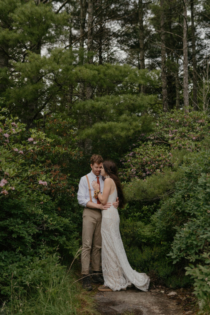 A couple stands amidst a forest with pines and rhododendron in their elopement clothes in North Carolina