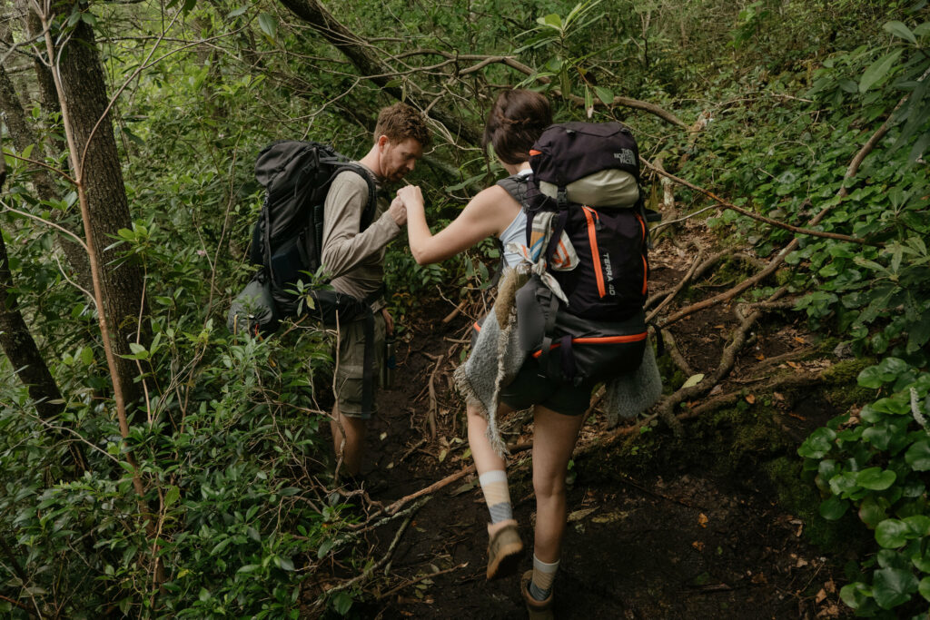 A couple is holding hands and hiking through a forest with a really muddy trail, backpacks full of gear for their camping elopement in North Carolina.