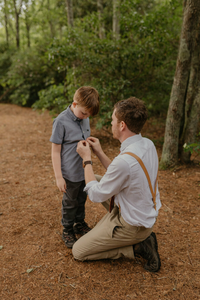 A groom is kneeling down helping button his sons shirt standing in nature. The Groom has wedding clothes on