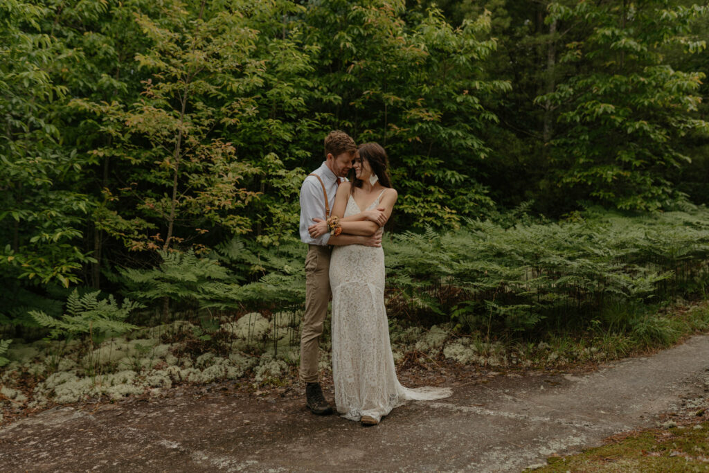 A couple is standing on an open rock face with trees and ferns and moss cuddled up facing each other during their forest elopement in North Carolina.