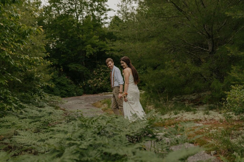 A couple walks by a grouping of tall ferns in a forest in North Carolina, looking at them and smiling. They are in their elopement clothes.