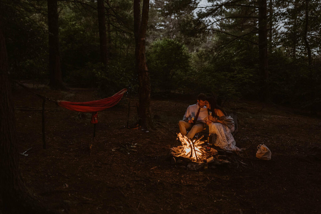 A couple sits in their elopement clothes snuggled up by the campfire and their hammock strung up beside them. They are in a forest in North Carolina and it is dusk.