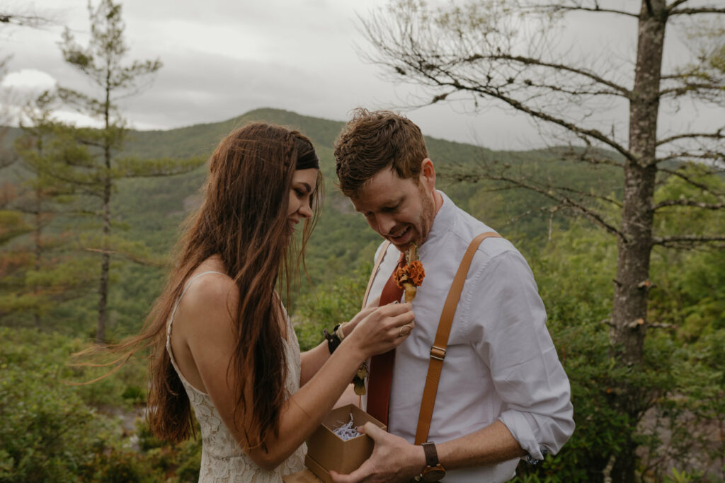 A couple is in a forest in North Carolina during their elopement. They are standing close looking at the boutonniere the bride designed and created.