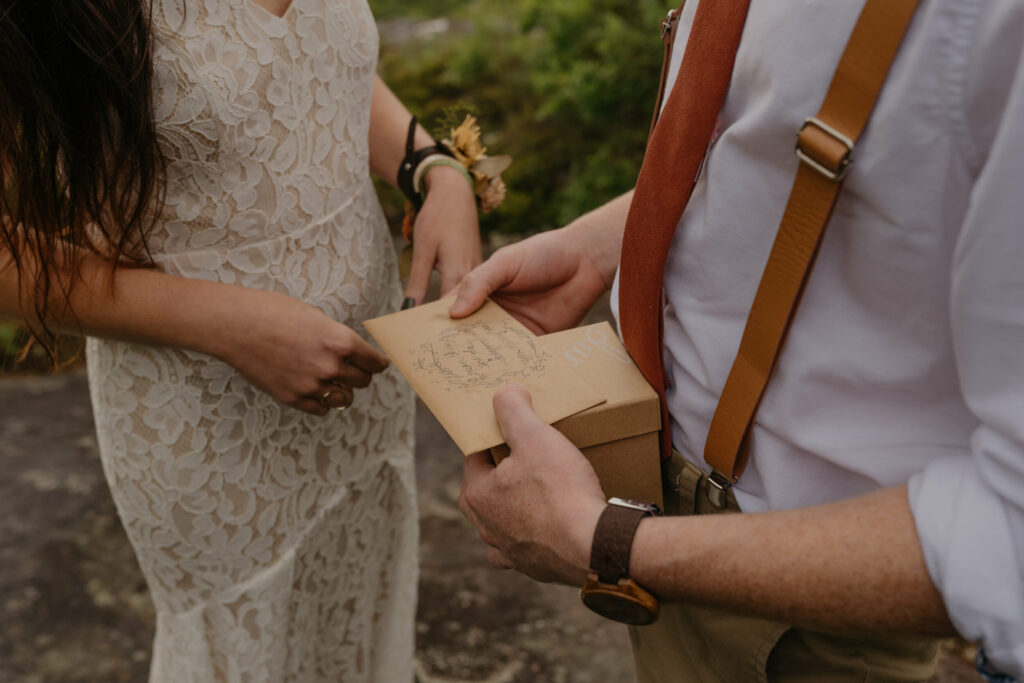 The hands of a couple in wedding clothes. The  groom is holding a letter envelope with writing on the outside and a box.