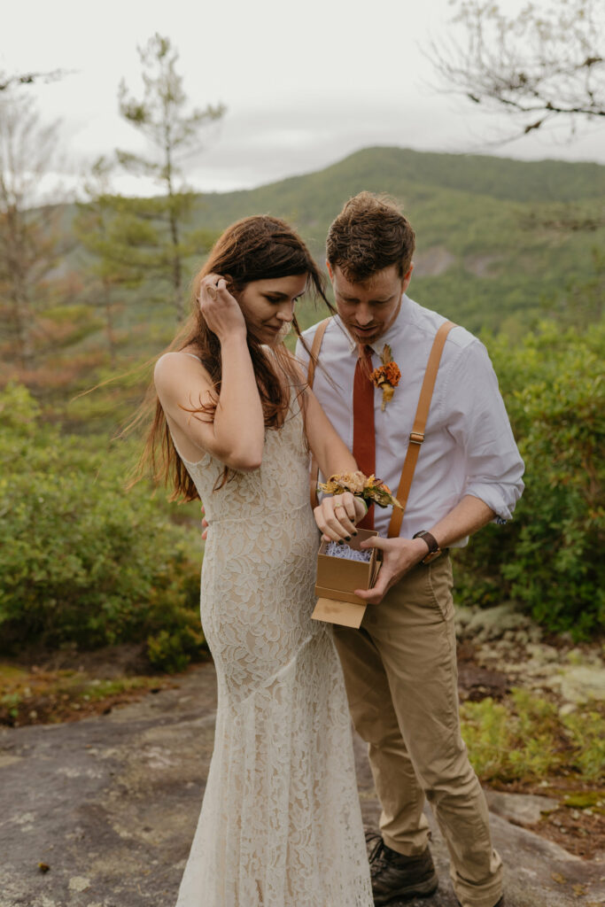 A couple is in a forest in North Carolina during their elopement. They are standing close looking at the floral wristlet the bride designed and created.