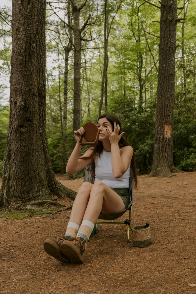 A bride is putting her makeup on while sitting in a camp chair in the the woods.