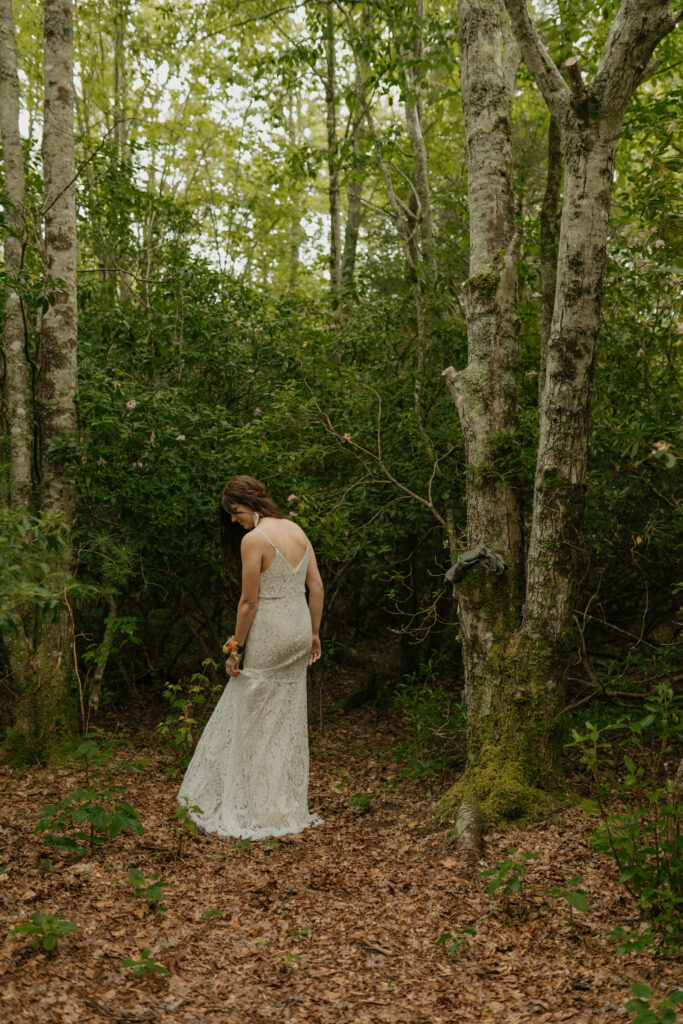 A bride is twirling around in the forest of North Carolina in her lacy elopement dress after getting dressed by a tree.