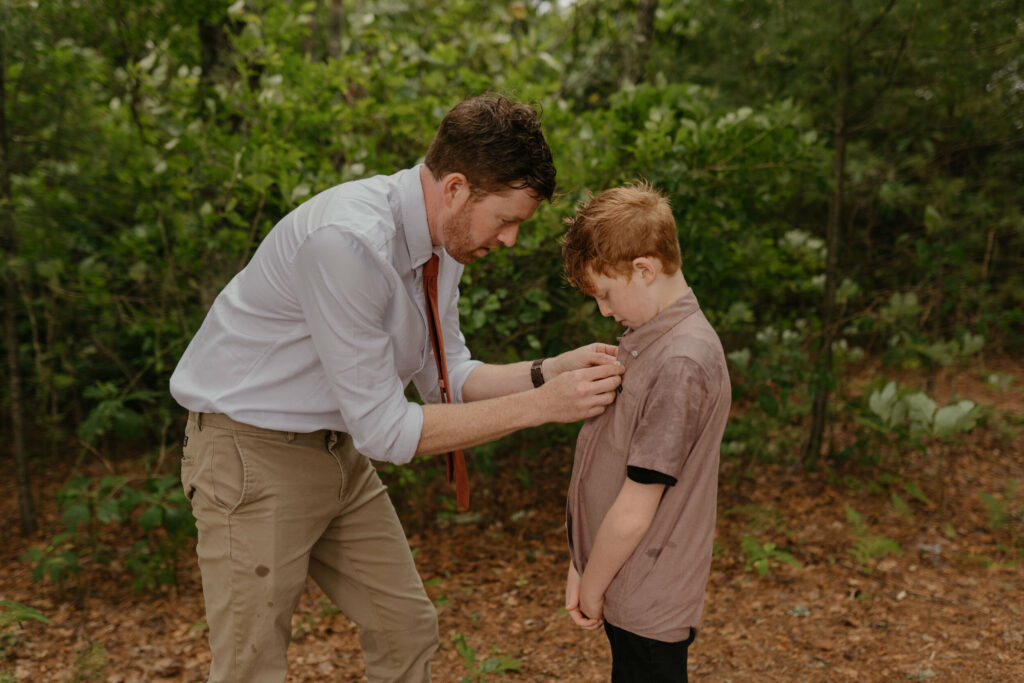 A groom is helping button his sons shirt in a forest in North Carolina during his elopement day.