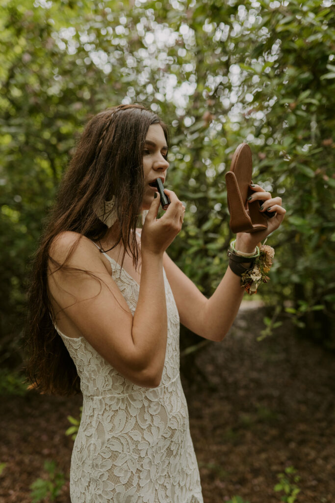 A bride is standing in the forest of North Carolina in her lacy elopement dress putting lipstick on in a small hand mirror.