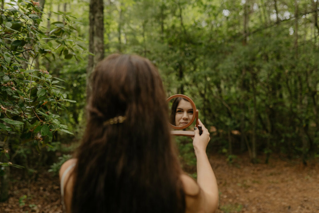 A bride is standing in the forest of North Carolina in her lacy elopement dress smiling at herself in a small hand mirror.