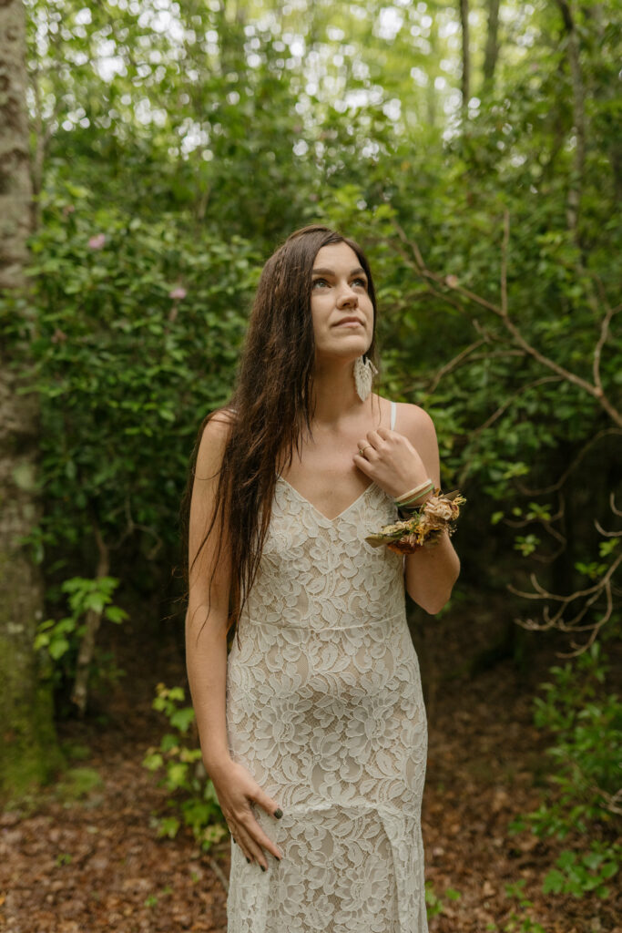 A bride is standing in the forest of North Carolina in her lacy elopement dress looking up at the sky.