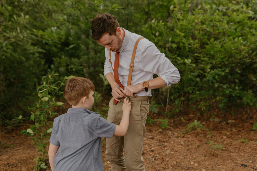 A son is helping tie his fathers clip in his suspenders in a forest in North Carolina during his fathers elopement day.