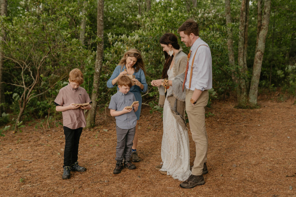 A couple is watching their kids and mother open gifts from them in nature in their wedding outfits.