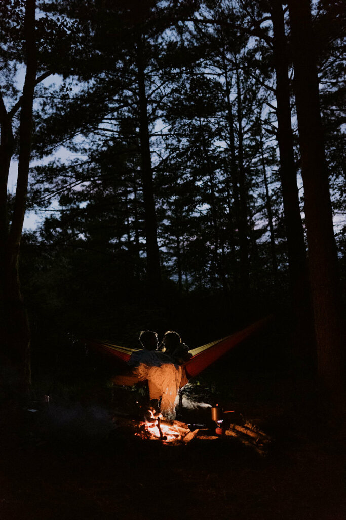 A couple sits in their elopement clothes in a hammock lit up by the campfire in front of them. It is sunset and the sky is blue through the pine forest behind them.