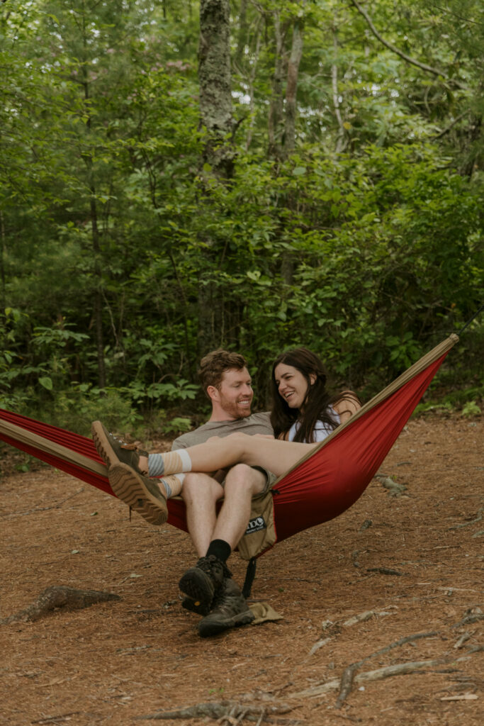 A couple sits cuddled in a hammock strung between two trees in the woods.