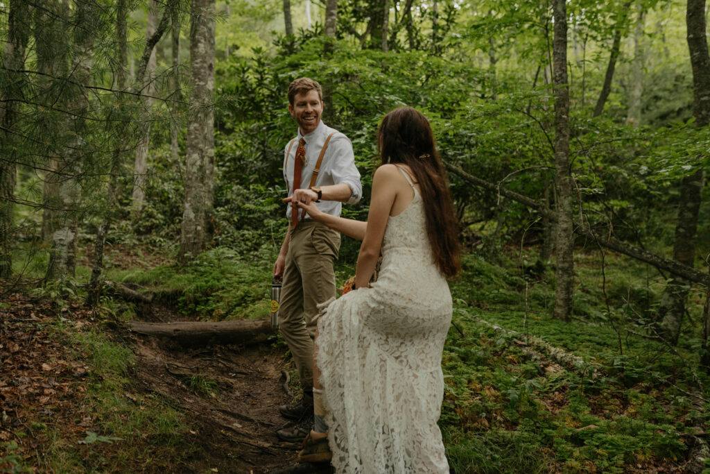 A couple is hiking through the forest in North Carolina during their elopement day. The groom is holding his brides hand helping her up a step with a smile on his face.