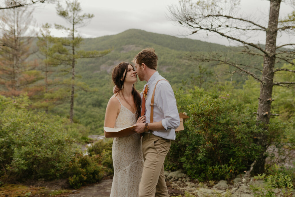 A couple is in a forest with a North Carolina mountain view in the background while they are looking at each other lovingly for their elopement day. The groom is holding a letter.