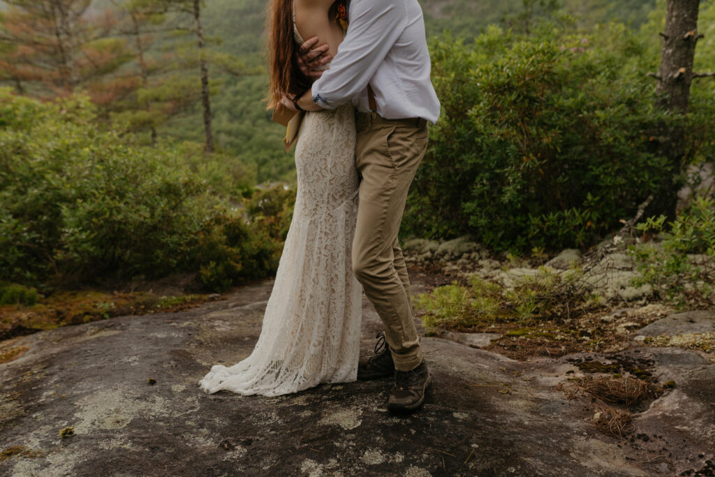 A couple is standing on a rock in a North Carolina forest hugging on their elopement day. You can only see the lower half of them and the details of the nature around them.