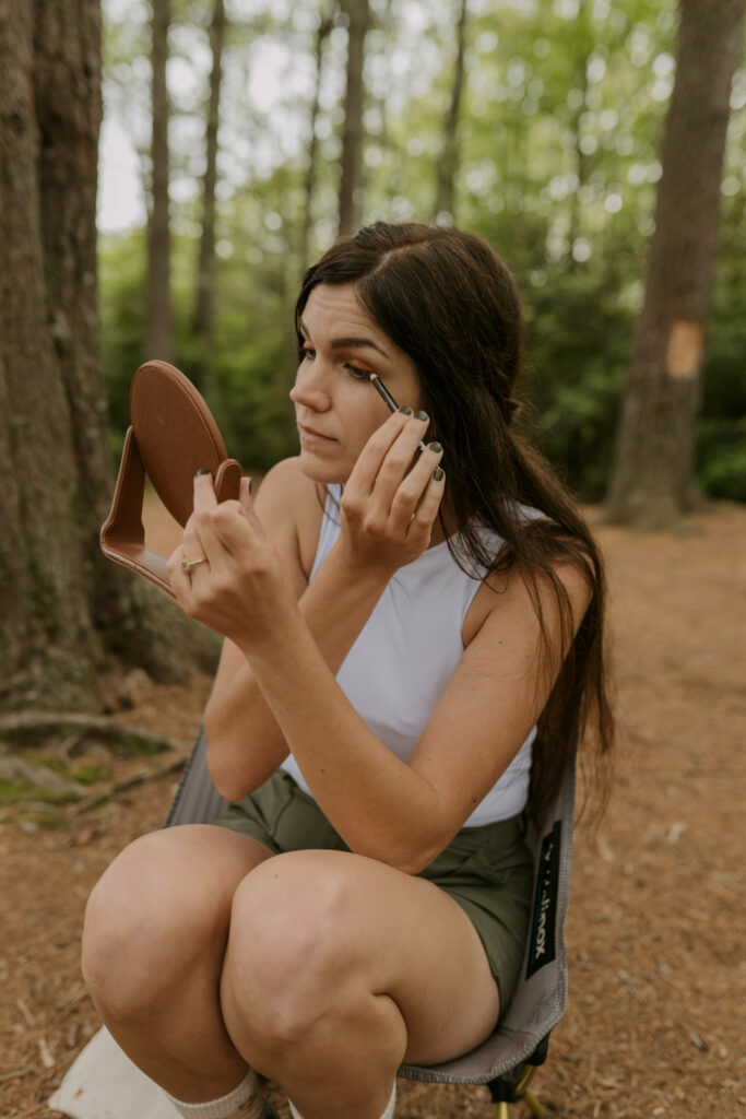 A bride is putting her makeup on while sitting in a camp chair in the woods.