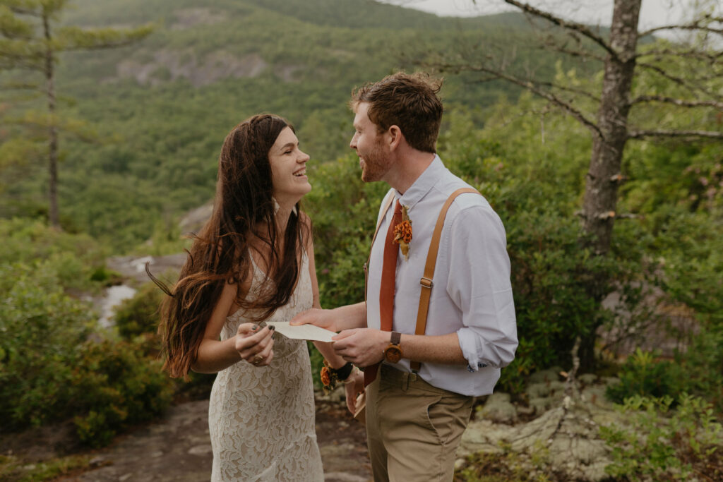 A couple is in a forest with a North Carolina mountain view in the background while they are looking at each other lovingly for their elopement day. The groom is holding a letter.