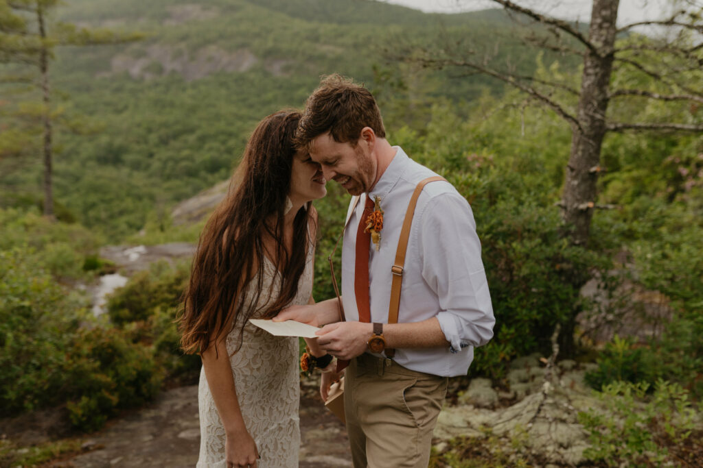 A couple is in a forest with a North Carolina mountain view in the background while they are forehead to forehead for their elopement day. The groom is holding and reading a letter.