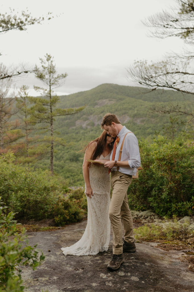 A couple is in a forest with a North Carolina mountain view in the background while the groom reads a letter his bride wrote him for their elopement day.