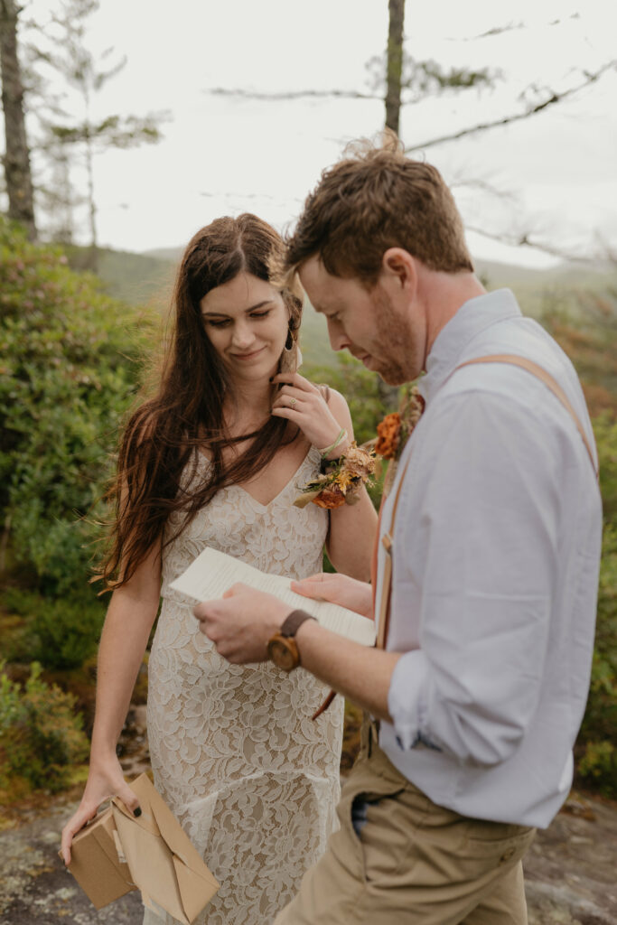 A couple is in a forest with a North Carolina during their elopement while the groom reads a letter and the bride is watching.