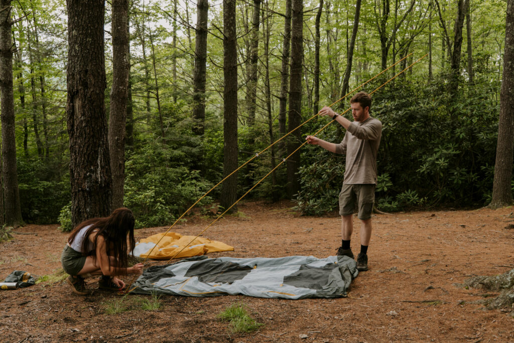 A couple is standing and putting together their tent in the woods.