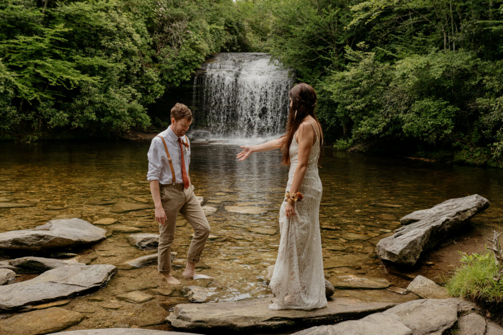 A bride is reach out to her groom on a rock in the river by a waterfall in North Carolina on their elopement day.