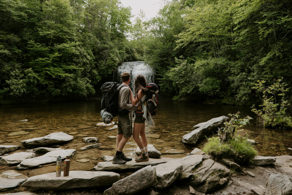A couple stands hugging with backpacks on looking at a waterfall surrounded by forest in North Carolina.