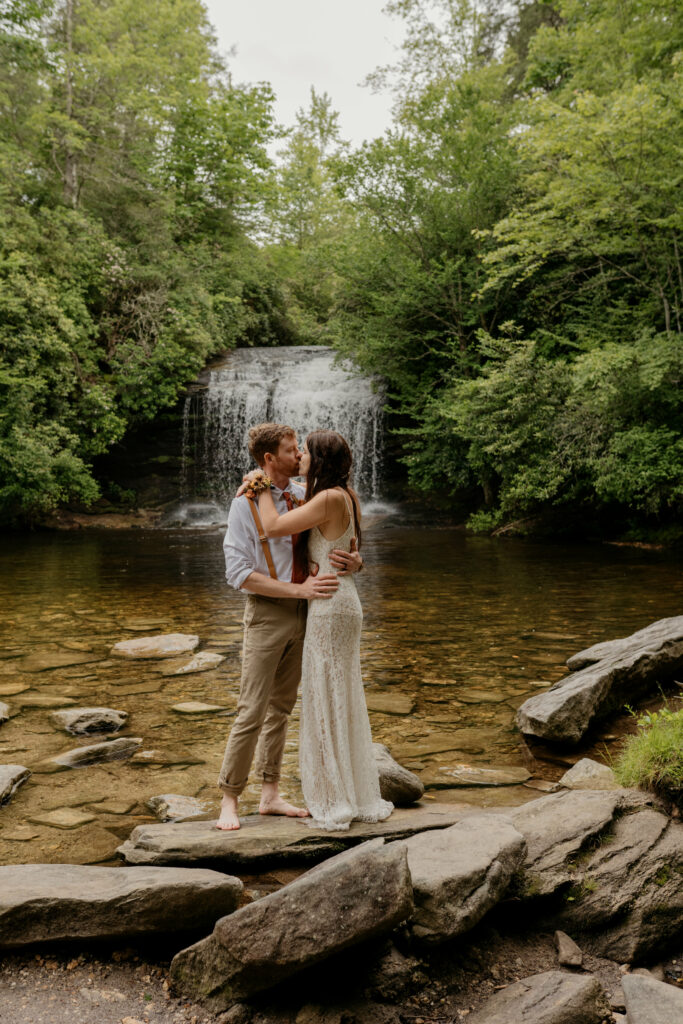 A couple is kissing on a rock in the river by a waterfall in North Carolina on their elopement day.