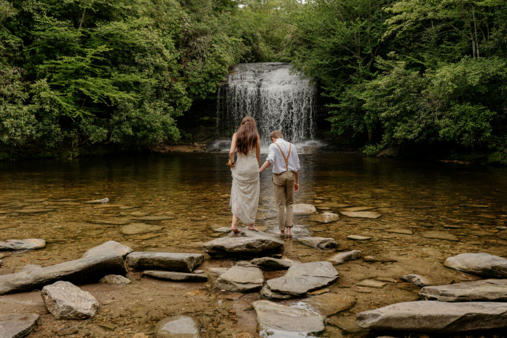 A couple is holding hands and walking barefoot in the river by a waterfall in North Carolina on their elopement day.