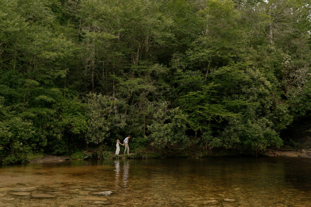 A couple is holding hands and walking barefoot in the forest by a river in North Carolina on their elopement day.